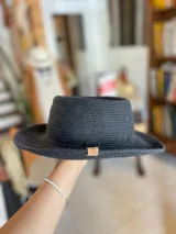 Sombrero negro de ala corta y copa plana, confeccionado en tejido trenzado de algodón con detalle de etiqueta de cuero en el lateral.
