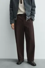 Pantalón de hombre color marrón oscuro, de corte balloon fit, con cintura con trabillas y cierre frontal de botón y cremallera. Presenta bolsillos delanteros y bolsillos traseros de vivo.