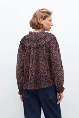 Blusa negra con estampado floral en tonos rosados y rojos, cuello con volados y mangas largas con puños elásticos.
