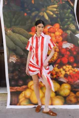 Vestido camisero corto a rayas verticales rojas y blancas con texto estampado. Tiene cuello camisero, mangas cortas y cierre frontal con botones.