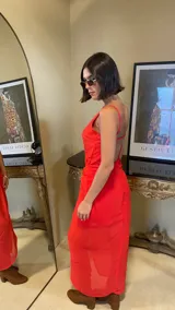 Vestido largo rojo con diseño asimétrico, escote redondo y detalle de nudo drapeado en la cintura que crea una abertura frontal, dejando ver una falda corta interior.