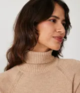 Suéter de tejido de punto color beige con cuello alto, mangas con corte raglán y puños y ruedo acanalados. Presenta un diseño clásico y cómodo con el ruedo trasero ligeramente más largo.