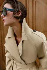 Trench corto de color beige, con cuello de solapa, manga larga con hombreras y trabillas con botones. Cuenta con cierre frontal cruzado con botones oscuros y cinturón ajustable del mismo tejido.