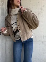 Campera bomber de gamuza color beige, con cierre frontal y detalle de ojales metálicos decorativos en la solapa. Presenta mangas largas con puños elásticos y un corte holgado tipo oversize.