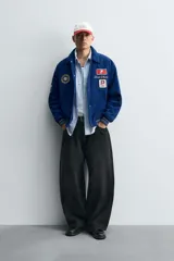 Cazadora estilo varsity confeccionada en pana de algodón azul, con interior ligeramente acolchado. Presenta cuello solapa, cierre frontal con botones a presión, bolsillos laterales de vivo y acabados en rib a rayas en puños y cintura. Incluye múltiples parches bordados decorativos en el frente.