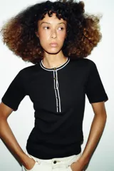 Camiseta de punto ajustada con cuello redondo y manga corta. Presenta detalles de vivos en color blanco a contraste en el cuello y la tapeta frontal, que cuenta con cierre de botones metálicos.