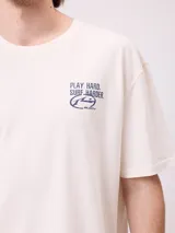 Remera de corte holgado color crema con estampado gráfico en la espalda que incluye el número 85, personajes animados y la frase Play Hard, Surf Harder.