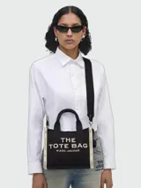 Cartera tipo tote bag de la marca Marc Jacobs, modelo The Small Tote, en color negro con detalles en blanco. Confeccionada en lona, presenta asas superiores y correa larga ajustable para llevar al hombro o cruzada. Cuenta con el texto "The Tote Bag" estampado en el frente.