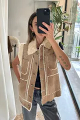 Chaleco de cuero genuino color tostado, con interior y detalles en corderito sintético color crema. Presenta cierre frontal con botones, cuello solapa y dos bolsillos frontales tipo parche.