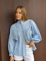 Camisa de algodón color celeste, con cuello mao, cierre frontal de botones y mangas largas abullonadas con puños elásticos. Presenta un diseño con costuras acolchadas en la parte superior delantera.