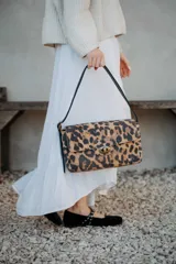 Cartera de hombro con diseño rectangular y solapa frontal, confeccionada en cuero con estampado animal print de leopardo. Cuenta con herrajes metálicos en tono bronce y una correa de hombro ajustable en color negro.