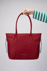 Bolso tipo tote bag color rojo con doble asa de hombro y detalles de tiras colgantes celestes en los laterales con cierre de cremallera.