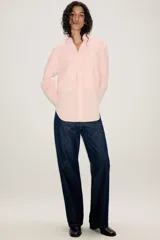 Camisa de corte oversize color rosa claro con diseño de rayas verticales sutiles. Presenta cuello solapa desmontable, cierre frontal con botones ocultos bajo solapa, mangas largas con puños y bajo asimétrico redondeado. Incluye dos bolsillos frontales amplios.