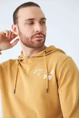 Buzo canguro color mostaza con capucha ajustable mediante cordones, bolsillo frontal tipo canguro y estampado gráfico en el pecho. Presenta puños y ruedo acanalados.