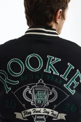 Campera estilo varsity con cuerpo negro y mangas en contraste color crema. Presenta múltiples parches y bordados decorativos en el frente y las mangas, cuello, puños y ruedo acanalados a rayas, y cierre frontal con botones a presión.