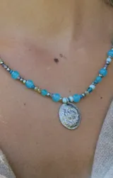 Gargantilla de cuentas de cristal en tonos tornasolados, violetas y oscuros, con dije central ovalado de metal plateado con diseño de escudo.