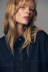 Camisa de denim color azul oscuro, con cuello solapa, manga larga y corte cropped. Presenta dos bolsillos de plastrón con tapa en el frente y cierre frontal con botones metálicos.