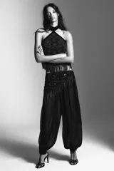 Pantalón bombacho negro semitransparente de tiro medio y cintura elástica. Tiene un pareo superpuesto ajustable con lazada lateral, decorado con abalorios, lentejuelas y flecos plateados. El bajo es elástico.