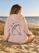 Canguro de felpa color crudo, con capucha, corte amplio, bolsillos delanteros y aberturas laterales en el dobladillo. Presenta un estampado en la espalda con diseño de arco y flecha rodeado de estrellas, y detalles gráficos en las mangas.
