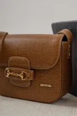 Cartera negra de cuero charolado con solapa y cierre de broche. Presenta un detalle metálico dorado en forma de freno de caballo en la parte frontal y una correa ajustable.