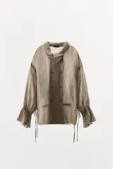 Camisa de lino color tostado con cuello subido, manga larga con puños elásticos y volantes, cintura ajustable con lazadas laterales y cierre frontal cruzado con doble botonadura.