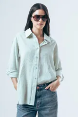 Camisa de corte oversize con diseño de rayas verticales finas en tonos claros, cuello clásico y cierre frontal con botones.