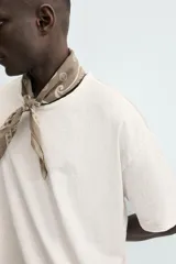 Camiseta de corte relajado (relaxed fit) color blanco roto, con cuello redondo y manga corta. Presenta un bordado sutil en el pecho con diseño floral en tonos claros.