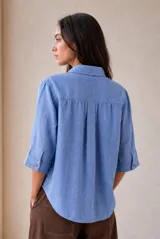 Blusa azul de lino con cuello camisero, escote en V y mangas tres cuartos con vuelta. Presenta un diseño holgado con dobladillo redondeado y un pequeño parche con logo bordado en el lateral inferior.