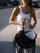 Bolso de hombro tipo hobo confeccionado en gamuza marrón, decorado con tachas metálicas plateadas y flecos largos en los laterales. Cuenta con cierre superior y correa ajustable para llevar al hombro o cruzado.