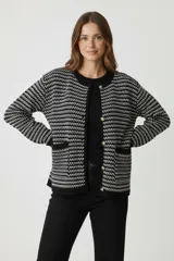 Cardigan de tejido de punto grueso, blanco y negro, con patrón geométrico en franjas horizontales. Es de corte holgado, sin cuello, con botones dorados frontales y dos bolsillos delanteros.