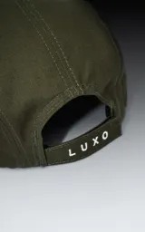 Gorra estilo 5 paneles, color verde militar en la corona y visera plana negra. Presenta un pequeño parche cuadrado negro con un logo blanco bordado en forma de tijeras o lazo.