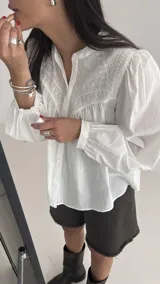 Camisa blanca de algodón con cuello mao, mangas largas con puños elásticos y cierre frontal de botones. Presenta un diseño con pliegues en la parte delantera y detalles bordados en el pecho.