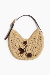 Cartera de hombro de paja trenzada color beige, con diseño semicircular, asa ajustable de cuero sintético marrón oscuro y detalles colgantes con nudo y apliques en forma de flor.