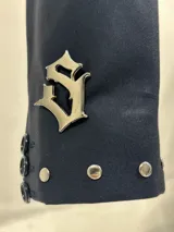 Blazer corto de color azul marino, con solapas decoradas con tachas metálicas plateadas y detalle de dije metálico en la manga. Presenta un corte estructurado y cierre frontal.