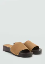 Sandalias de suela de madera color marrón oscuro, con una única tira ancha en piel de serraje color tostado decorada con tachas doradas a lo largo de los bordes.