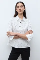 Blusa blanca con estampado de pequeños lunares negros, cuello camisero y botones frontales oscuros. Mangas tres cuartos con ligero vuelo en el puño.