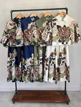Conjunto de dos piezas confeccionado en viscosa con estampado floral en tonos marrones y verdes sobre base blanca. Incluye una blusa tipo camisa corta con cuello solapa, cierre frontal de botones y mangas abullonadas con diseño globo, y un pantalón tipo culotte de tiro alto, amplio y fluido con pinzas en la cintura.