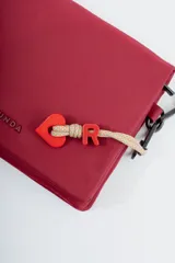 Bandolera de diseño rectangular en color verde oliva, con solapa frontal y logo de la marca grabado. Incluye una correa larga ajustable con el nombre de la marca tejido y un charm decorativo con forma de corazón y letra R en color rojo.