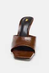 Zapato destalonado tipo mule de piel con acabado charol marrón, tira ancha delantera y tacón alto fino en contraste de materiales. La punta es cuadrada.