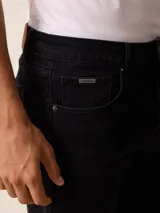 Jean de corte recto y tiro medio, confeccionado en denim de color negro con costuras a tono. Cuenta con cierre frontal con botón metálico, cinco bolsillos tradicionales y pasacintos en la cintura.
