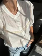 Camisa de estilo romántico color blanco, confeccionada en tejido liviano con textura de rayas verticales. Presenta mangas al codo con frunces en los hombros, cuello redondo y cierre frontal con botones. Incluye delicados detalles de puntilla de algodón en el pecho y en los bordes de las mangas.