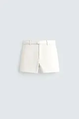Shorts de algodón color blanco roto, de corte regular fit, con cintura ajustable mediante cinturón del mismo tejido y hebilla metálica. Presenta bolsillos delanteros y bolsillos traseros de vivo.