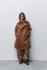 Trench clásico color marrón, de silueta relajada, confeccionado en tejido liviano, con cuello de solapa, doble abotonadura frontal y mangas largas con presillas regulables en los puños.