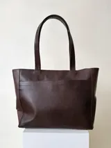 Cartera tipo tote de cuero graneado color marrón oscuro, con textura uniforme y suave al tacto. Tiene un bolsillo frontal con broche imantado, un bolsillo trasero con cierre metálico y cierre principal también metálico. En el interior, está forrada y cuenta con tres bolsillos: uno con cierre, uno tipo plaqué para celular y un bolsillo grande tipo sobre con solapa y botón. Las asas son de 55 cm de largo.
