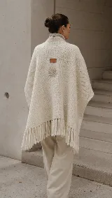 Poncho de lana natural en color crudo, con silueta envolvente y diseño arquitectónico. Presenta flecos largos en el ruedo, un detalle bordado en lino en la espalda y una etiqueta de cuero distintiva.