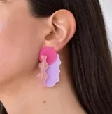 Par de aros colgantes con diseño abstracto en forma de silueta femenina, con círculos fucsia en la parte superior, siluetas color cobre con glitter en el centro y formas irregulares color lila en la parte inferior.