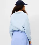 Buzo de algodón color celeste con cuello polo blanco en contraste. Presenta un diseño de estilo universitario con estampado frontal con relieve y mangas largas con puños acanalados.