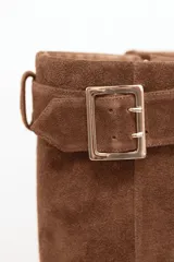 Bota de caña alta confeccionada en cuero gamuza color marrón, con diseño de solapa superior y detalle de hebilla metálica lateral. Presenta punta fina y suela plana.