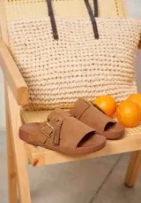 Sandalias de gamuza color marrón, con diseño de una sola tira ancha en el empeine decorada con una hebilla metálica dorada y una borla de flecos colgantes.