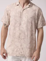 Camisa de manga corta color beige con estampado de hojas en tonos marrones. Confeccionada en viscosa, tiene cuello tipo solapa y cierre frontal de botones.
